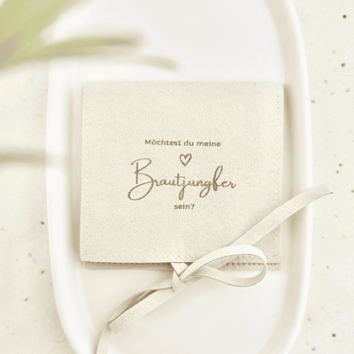 Beigefarbener Schmuckbeutel mit Schleifenband und goldener Aufschrift „Möchtest du meine Brautjungfer sein?“ auf einem weißen Teller, Geschenkverpackung für Schmuck zur Brautjungfer-Frage.