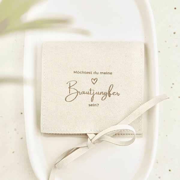 Beigefarbener Schmuckbeutel mit Schleifenband und goldener Aufschrift „Möchtest du meine Brautjungfer sein?“ auf einem weißen Teller, Geschenkverpackung für Schmuck zur Brautjungfer-Frage.