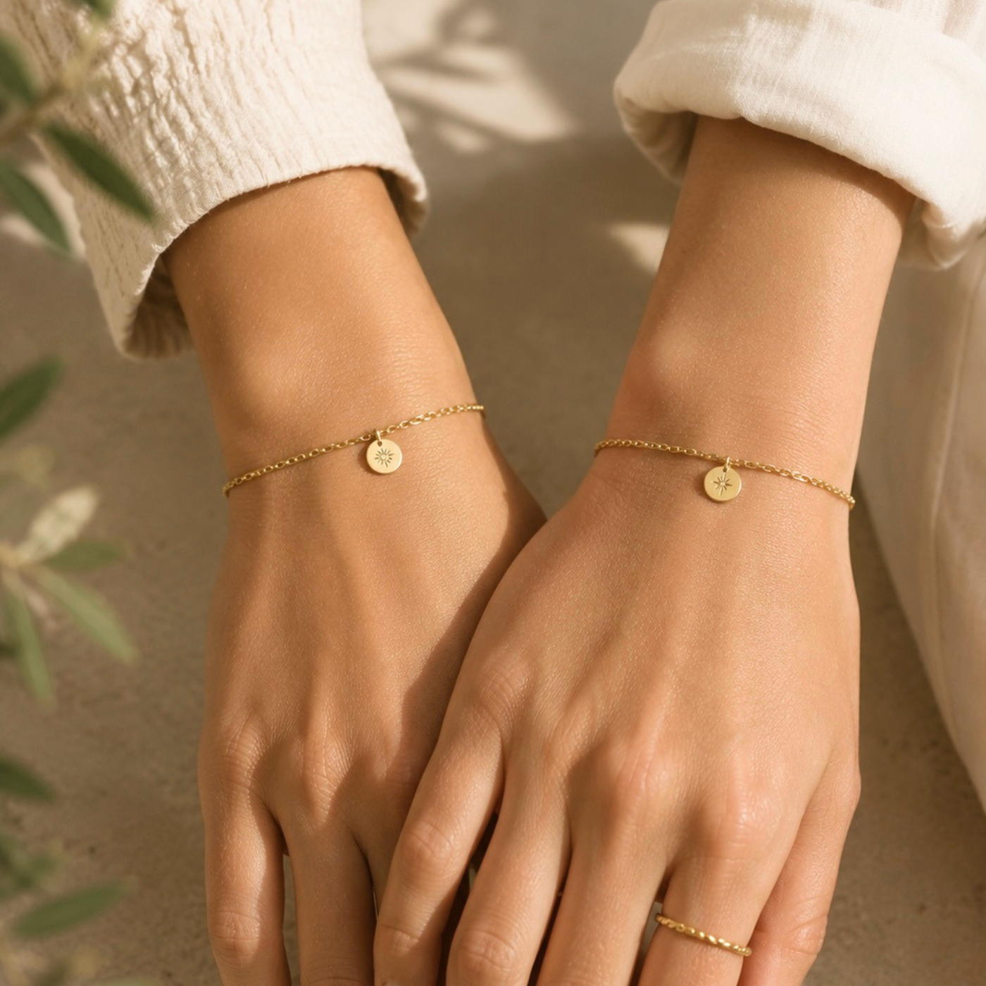 Zwei Hände mit filigranen goldfarbenen Permanent Armbändern und kleinen Anhängern in natürlicher Umgebung. Permanent Schmuck Termin im Studio in Wuppertal.