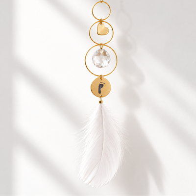 Personalisierter Sonnenfänger in Gold mit Herzanhänger, Kristallkugel, Baby-Fußabdruck-Gravur und weißer Feder vor hellem Hintergrund.